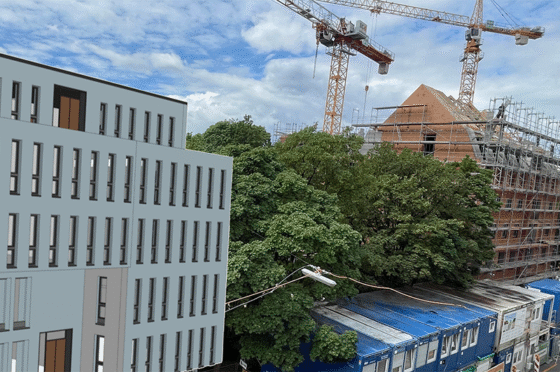 Moderner Gebäudekomplex neben einer Baustelle mit Krananlagen und eingerüstetem Ziegelbau, teilweise von Bäumen verdeckt, unter bewölktem Himmel.