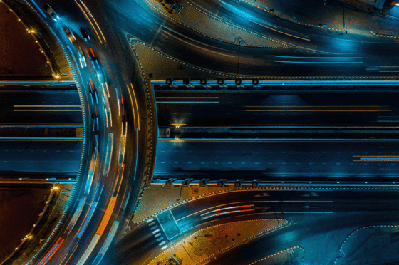 Nächtliche Luftaufnahme eines beleuchteten Autobahnkreuzes mit mehreren Fahrspuren und Lichtspuren fahrender Fahrzeuge. Die sich kreuzenden Verkehrswege symbolisieren Verbindungen, Abhängigkeiten und Netzwerke zwischen verschiedenen Elementen.