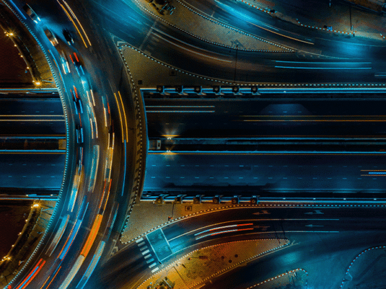 Nächtliche Luftaufnahme eines beleuchteten Autobahnkreuzes mit mehreren Fahrspuren und Lichtspuren fahrender Fahrzeuge. Die sich kreuzenden Verkehrswege symbolisieren Verbindungen, Abhängigkeiten und Netzwerke zwischen verschiedenen Elementen.