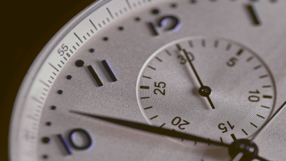 Nahaufnahme des Zifferblatts einer analogen Armbanduhr. Sichtbar sind die Zahlen 10, 11 und 12 auf dem äußeren Zifferblatt sowie ein kleines Hilfszifferblatt mit feinen Skalenmarkierungen. Die silberfarbene, strukturierte Oberfläche und die dunklen Zeiger sind scharf fokussiert, während die Bildränder durch die geringe Schärfentiefe weich verschwimmen. Warmes Licht betont die feinen Details und die hochwertige Verarbeitung der Uhr.